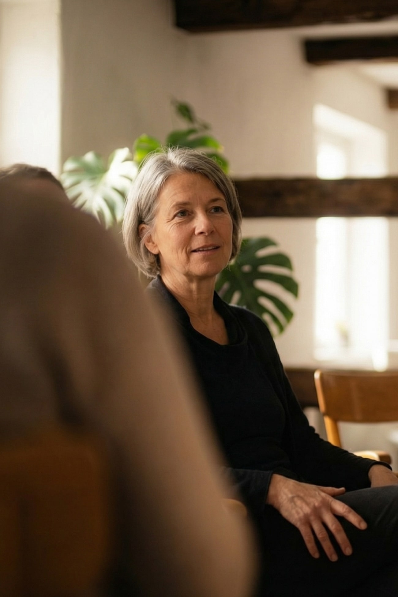 Eine Frau mit grauen Haaren sitzt in einem Raum und hört aufmerksam zu, umgeben von Pflanzen und Holzdetails.