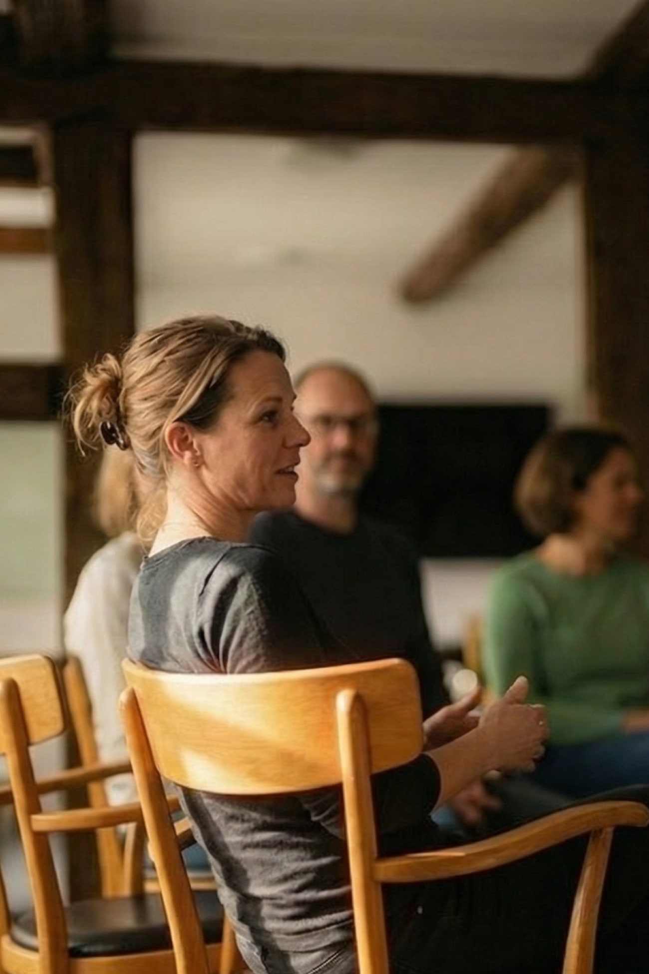 Eine Gruppe von Menschen sitzt in einem Raum, eine Frau spricht, während andere zuhören. Holzstühle im Vordergrund.