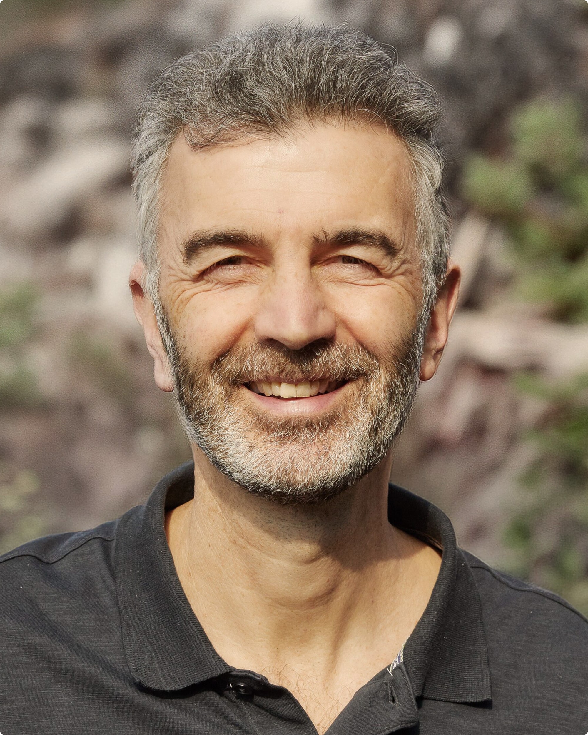 Porträt eines lächelnden Mannes mit grauen Haaren und Bart, im Freien vor unscharfem Hintergrund.