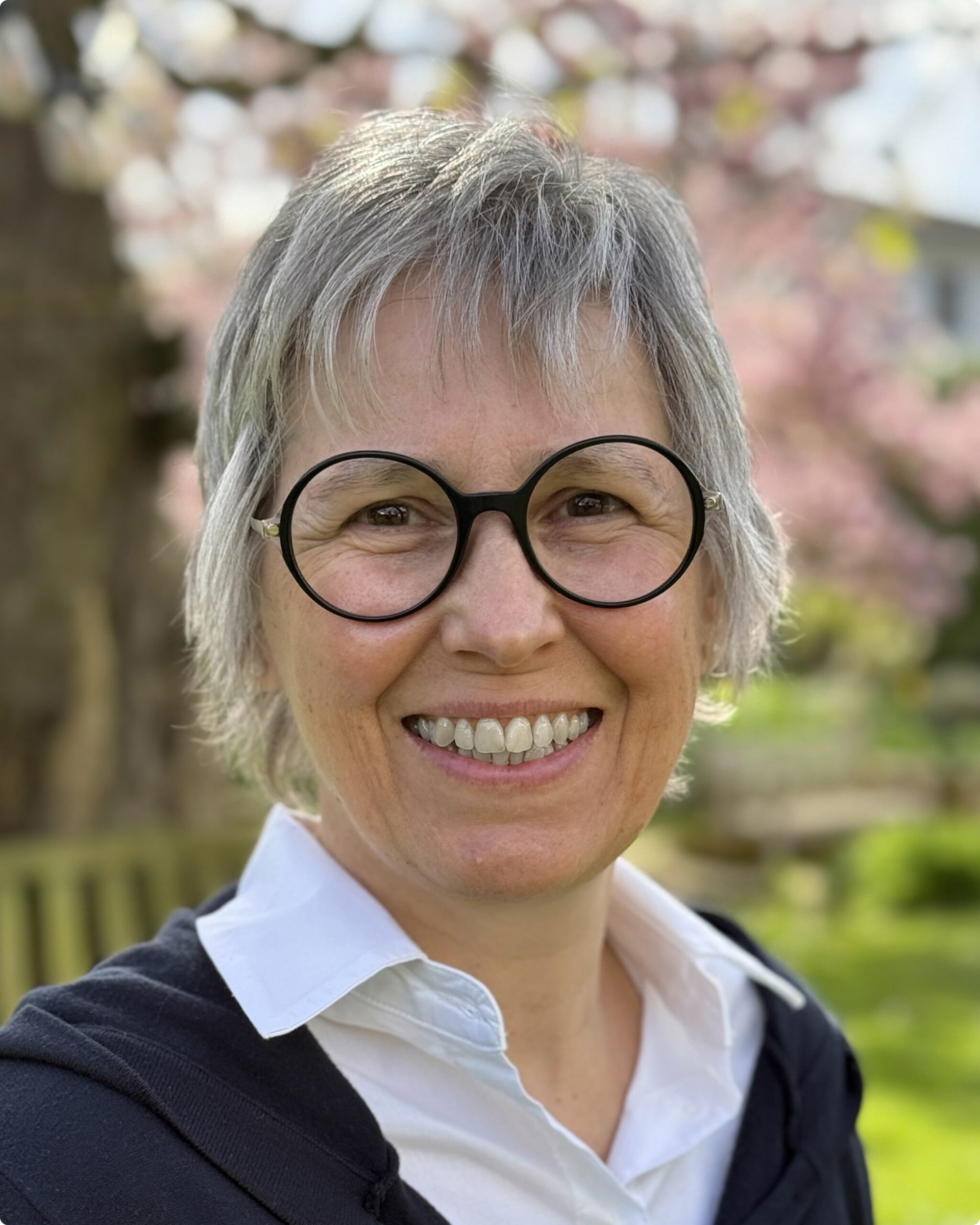 Porträt von Susanne Korpiun mit grauen Haaren und runden Brillen, lächelnd vor blühenden Bäumen im Hintergrund.