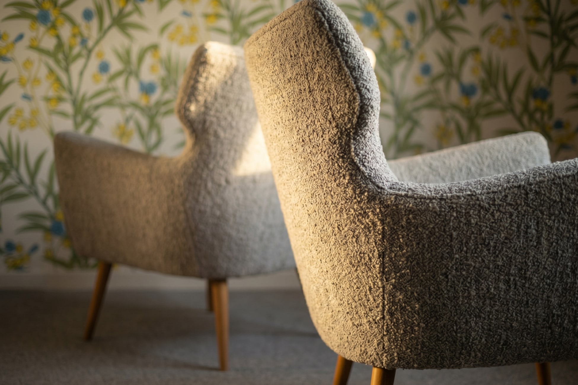 Zwei graue Sessel mit Holzbeinen vor einer blumigen Tapete in sanftem Licht.